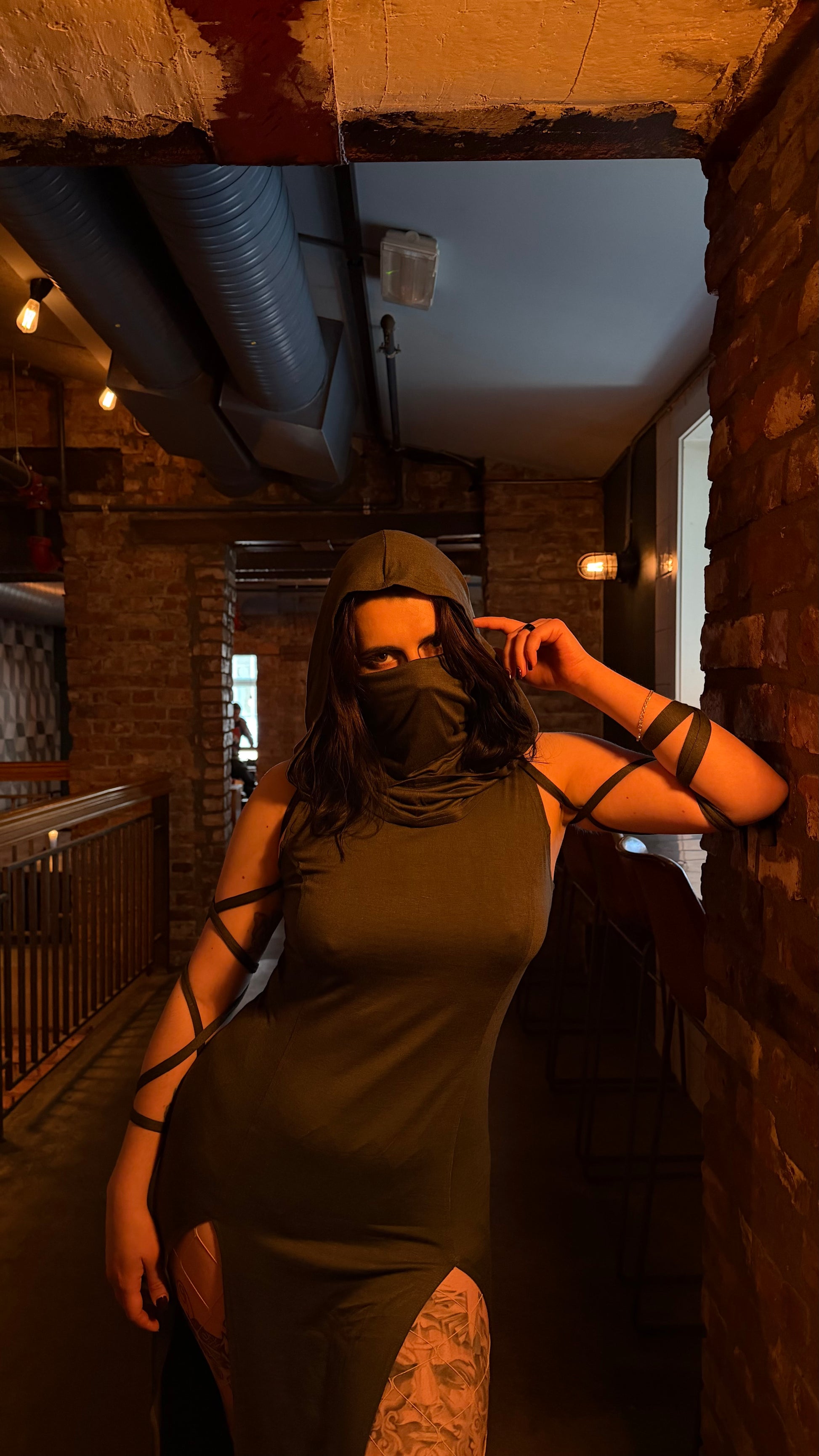 Person wearing a green dress with black straps in an indoor setting with brick walls and exposed pipes.